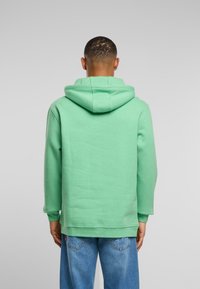 Mężczyzna w miętowej bluzie z kapturem i niebieskich dżinsach stoi plecami do jasnoszarego tła.