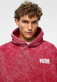 Rode hoodie van zacht materiaal, met een grote voorkant zak en witte tekst "ESCAPE FROM THE POLIZIE" op de linkerkant van de borst.