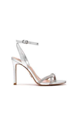 Sandalo argento con tacco alto, cinturino alla caviglia e fasce incrociate sulle dita decorate con strass, mostrato di lato su sfondo bianco.