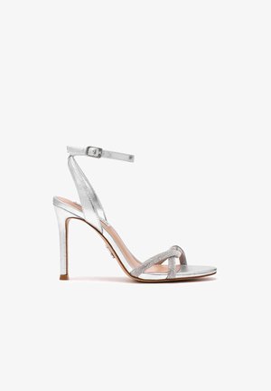 Sandalo argento con tacco alto, cinturino alla caviglia e fasce incrociate sulle dita decorate con strass, mostrato di lato su sfondo bianco.