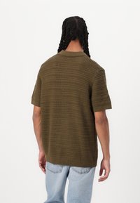 Chemise tricotée à manches courtes de couleur vert olive avec des rayures texturées, un col et un ourlet côtelés. Portée avec un jean bleu clair à coupe ample.