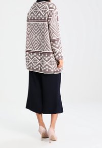 Lungo cardigan lavorato a maglia in crema e borgogna con motivi geometrici, abbinato a culotte blu scuro e décolleté beige.