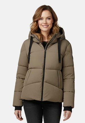 Vrouw met schouderlang golvend haar draagt een taupekleurige pufferjas met capuchon, zwarte trekkoorden en zwarte broek, staat en glimlacht.