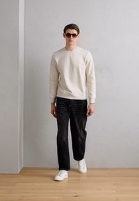 Felpa beige chiaro con design minimale, jeans neri e sneakers bianche. L'individuo indossa occhiali da sole scuri. Sfondo semplice.