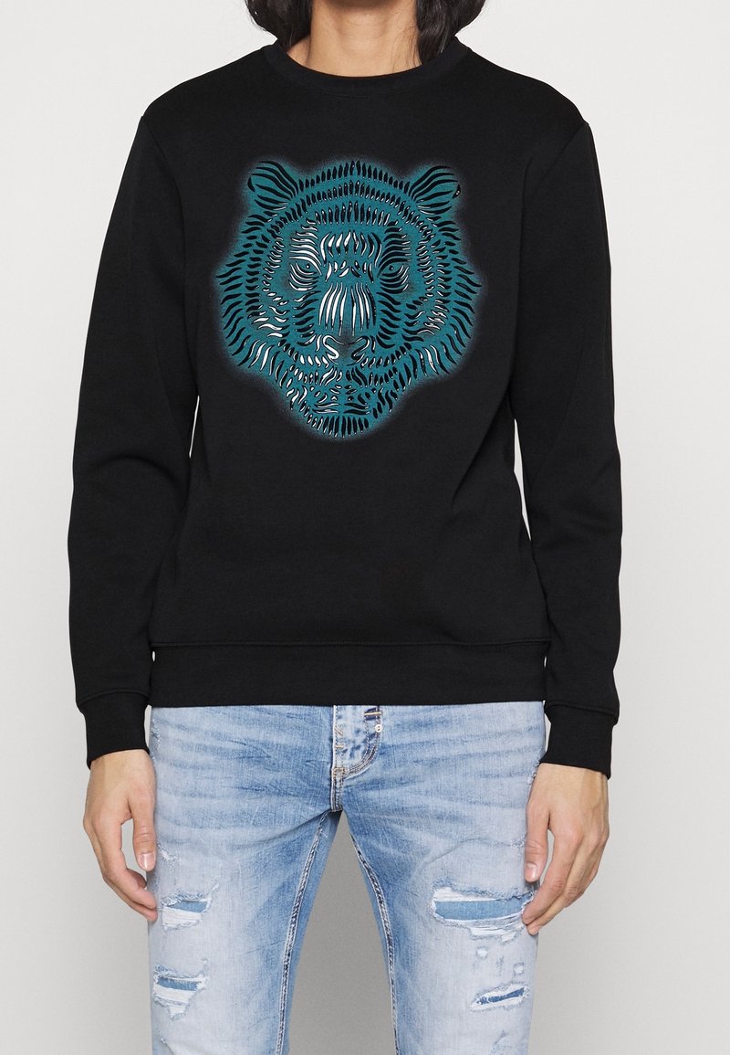 Sweat-shirt noir avec un graphisme stylisé de visage de tigre bleu et blanc, porté avec un jean déchiré bleu clair.