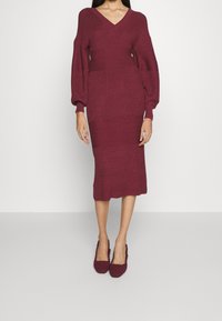 Abito in maglia bordeaux con scollatura a V e lunghe maniche a palloncino; presenta motivi a coste orizzontali. Abbinato a scarpe slip-on testurizzate bordeaux.