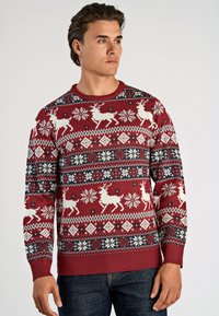 Mann in rotem Weihnachtspullover mit weißen Rentieren, Schneeflocken und geometrischen Mustern, kombiniert mit dunkelblauen Jeans, schaut nach rechts.