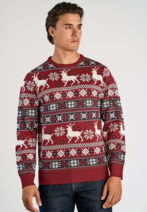 Mann in rotem Weihnachtspullover mit weißen Rentieren, Schneeflocken und geometrischen Mustern, kombiniert mit dunkelblauen Jeans, schaut nach rechts.