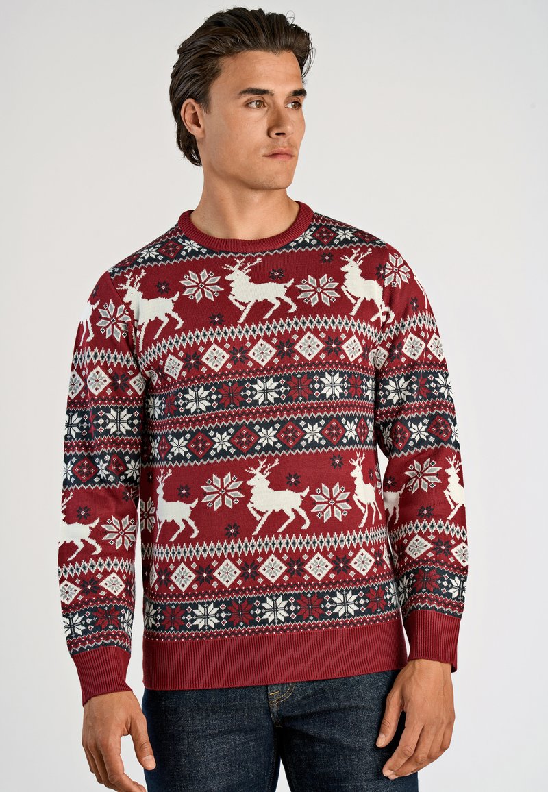 Mann in rotem Weihnachtspullover mit weißen Rentieren, Schneeflocken und geometrischen Mustern, kombiniert mit dunkelblauen Jeans, schaut nach rechts.