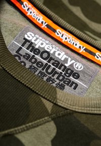 Zelená maskáčová mikina s žebrovaným límcem, na které je našitá černá a oranžová nálepka "Superdry" a šedá pečeť o údržbě uvnitř.