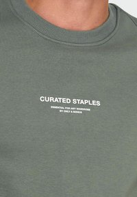 Grøn sweatshirt med ribbet halsudskæring og trykt tekst "CURATED STAPLES" i hvid. Casual design og stof i bomuldsblanding.