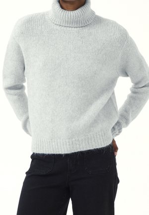 Osoba ubrana w jasnoszary, puszysty sweter z golfem i czarne spodnie, stojąca z rękami za plecami na białym tle.