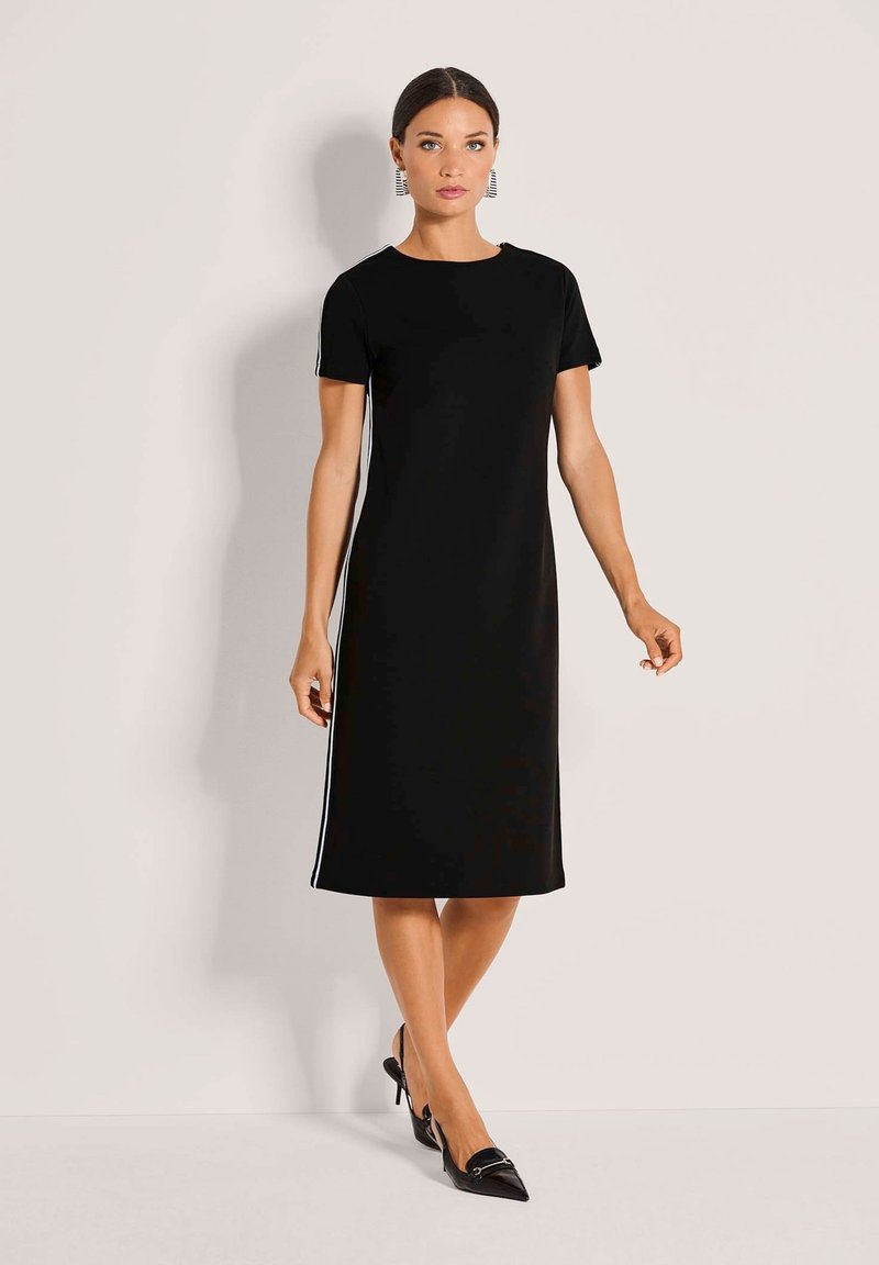 Schwarzes kurzärmliges Kleid mit geradem Schnitt, weißen Seitenstreifen und Rundhalsausschnitt, kombiniert mit schwarzen spitzen Schuhen.