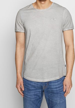 Homme portant un t-shirt gris clair à manches courtes et un jean bleu, debout avec les bras détendus le long du corps devant un fond uni.