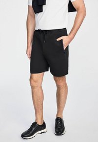 Czarne szorty z elastycznym pasem i sznurkiem, z bocznymi kieszeniami i gładkim wykończeniem materiału. W połączeniu z czarnymi butami sportowymi.