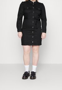Robe chemise en denim noir avec des manches longues, deux poches poitrine, fermeture à boutons, taille ceinturée et une silhouette droite, portée avec des chaussettes blanches et des chaussures noires.