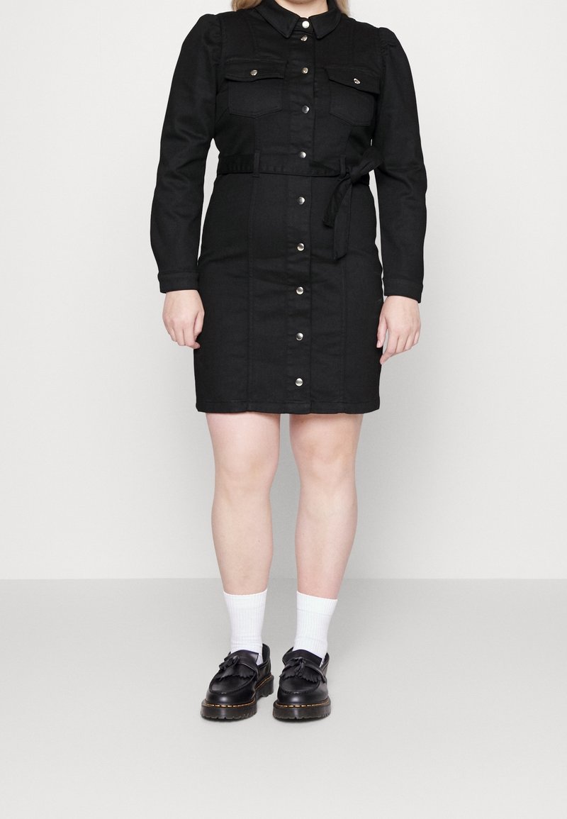 Robe chemise en denim noir avec des manches longues, deux poches poitrine, fermeture à boutons, taille ceinturée et une silhouette droite, portée avec des chaussettes blanches et des chaussures noires.