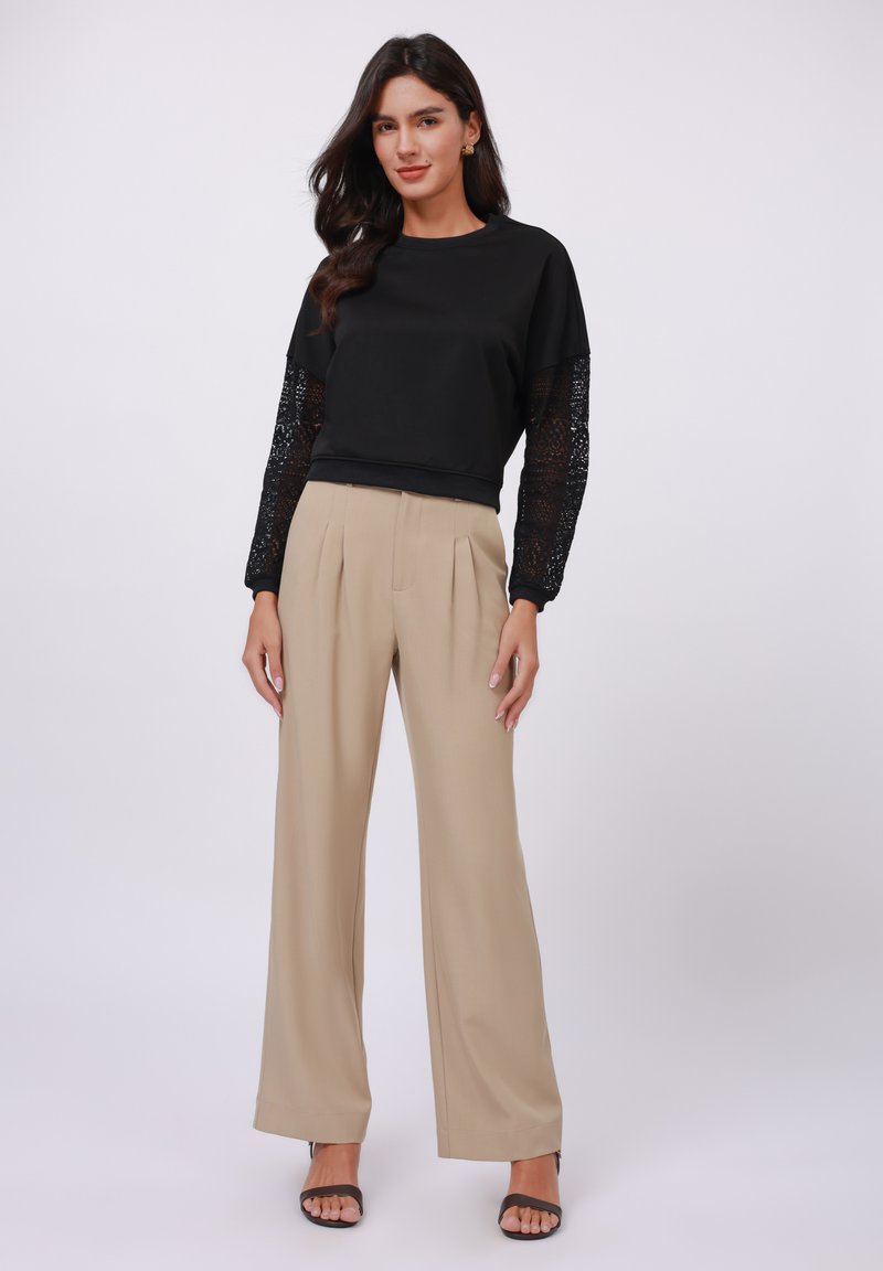 Schwarzes Langarmoberteil mit Spitze an den Ärmeln, kombiniert mit hoch taillierten beigen weiten Hosen und schwarzen offenen High Heels.