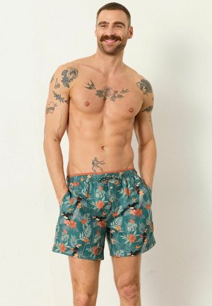 Lächelnder, oberkörperfreier Mann mit Schnurrbart und Tätowierungen auf Armen und Brust, bekleidet mit grünen, geblümten Badeshorts mit Taschen und Kordelzug.