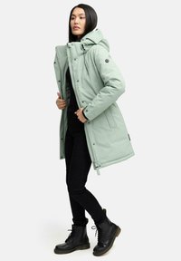 Parka rembourrée vert clair avec capuche, dotée d'une fermeture éclair et de boutons-pression, de poches latérales et d'une coupe décontractée. Bottes de combat noires.