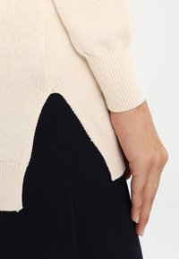 Hellbeige gestrickter Pullover mit gerippten Ärmeln, der an der Seite einen Schlitz für zusätzliche Details aufweist. Die Textur ist weich und leicht durchscheinend.