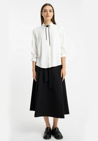 Biała bluzka z czarnym detalem w postaci krawata, z rękawami 3/4 i marszczonymi mankietami, zestawiona z długą czarną spódnicą. Modelka nosi czarne buty.