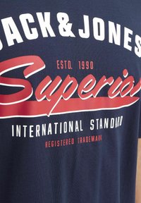 Marineblauwe katoenen t-shirt met witte en rode grafische tekst. Bevat "JACK & JONES," "SUPERIOR," en "INTERNATIONAL STANDARD." Zachte textuur.