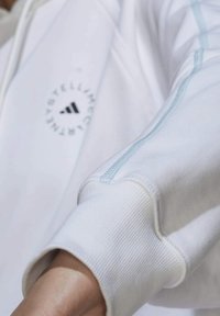 Vit hoodie med blå accenter, ribbade cuffs och tryckt logotyp. Tyget verkar mjukt med en slät textur och har kontrasterande sömmar.