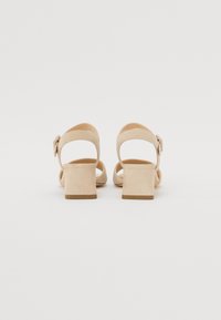 Beige suede heeled sandals with an open back, three straps, and a rectangular heel. Features a small bow detail on the ankle strap.
