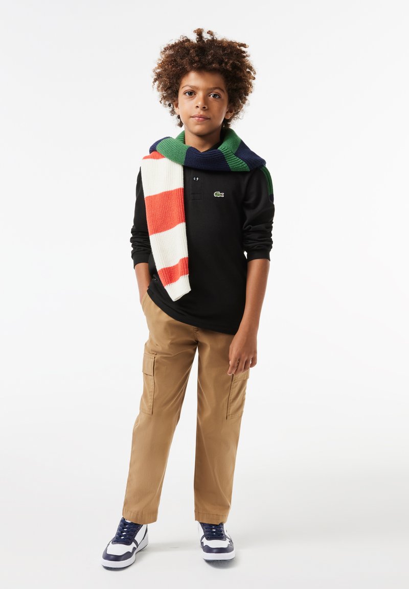 Black long-sleeve polo shirt with green logo, tan cargo pants, and white-blue sneakers. A striped knitted sweater rests over the shoulders.