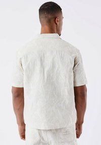 Camisa de manga corta en beige claro, con tejido texturizado y un patrón sutil. Presenta un cuello clásico y un dobladillo recto.