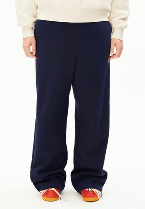 Person trägt lockere dunkelblaue Hose und rote Sneakers mit gelben Sohlen, steht mit entspannten Händen neben dem Körper vor weißem Hintergrund.