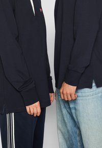 Two people stand side by side wearing navy long-sleeve shirts. The shirts have a short collar and slight side slits at the hem.