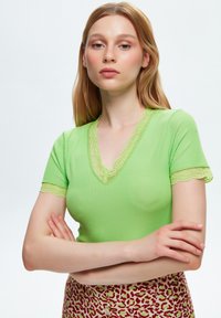 Maglietta corta a maniche corte di un verde chiaro a coste con scollatura a V e bordi in pizzo. Il modello la indossa con una gonna stampata, braccia conserte.