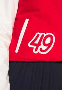 Veste varsity rouge et blanche avec le numéro 49 brodé, associée à une jupe plissée bleu marine.