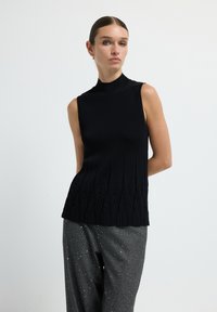 Vrouw met gelikt haar in een mouwloze zwarte tricot top en grijs gespikkelde broek, staand tegen een eenvoudige witte achtergrond.