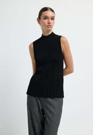 Vrouw met gelikt haar in een mouwloze zwarte tricot top en grijs gespikkelde broek, staand tegen een eenvoudige witte achtergrond.