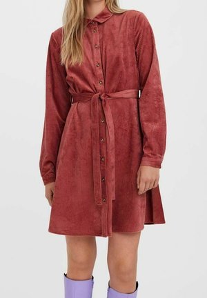 Kvinde iført en langærmet, knappet, rustrød cordkjole med bælte bundet i taljen og lilla støvler, der går til midt på læggen.