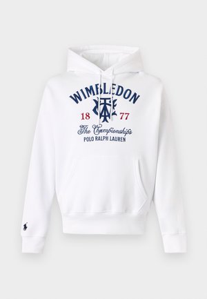 Άνδρας με λευκή μπλούζα με κουκούλα Wimbledon Polo Ralph Lauren, μπεζ cargo παντελόνια και μπεζ μοκασίνια, που στέκεται μπροστά σε ένα ομοιόμορφο ανοιχτό γκρι φόντο.