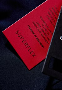 Rotes Produktlabel mit schwarzem Text, der "SUPERFLEX" hervorhebt. Die Textdetails konzentrieren sich auf Komfort und Bewegungsfreiheit, eingebettet auf einem dunklen Stoffhintergrund.