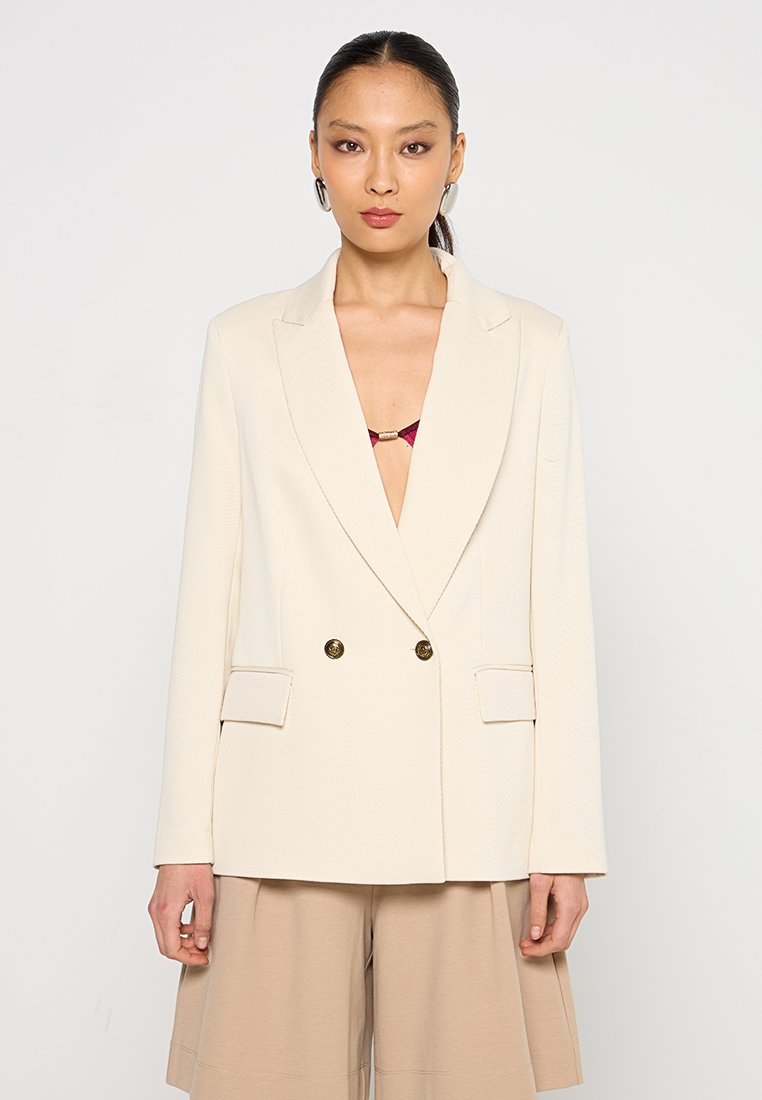 Blazer doppiopetto color crema in tessuto testurizzato, con rever a punta e bottoni dorati. Tasche frontali, abbinato a pantaloni corti beige.