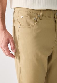 Beige puuvillapantsit suoran leikkauksen kanssa, joissa on etutaskut, hopeanväriset yksityiskohdat ja näkyvät ompelusaumat.