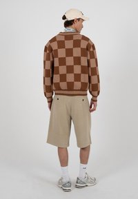 Homme de dos portant un pull à carreaux marron, un short beige, des chaussettes blanches, des baskets argentées et une casquette beige clair sur fond uni.