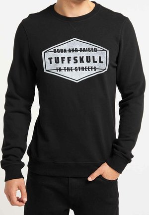 Mężczyzna ubrany w czarną bluzę z długim rękawem z białym logo w kształcie tarczy z napisem „Born and raised TUFFSKULL in the streets” na piersi.