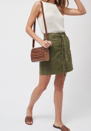 Femme portant un haut en maille blanc sans manches, une jupe ceinturée vert olive, des sandales tressées marron et un sac bandoulière en bois à lattes.