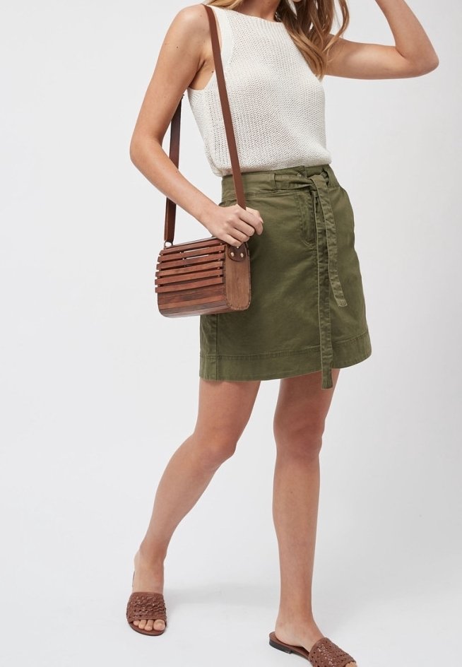 Femme portant un haut en maille blanc sans manches, une jupe ceinturée vert olive, des sandales tressées marron et un sac bandoulière en bois à lattes.