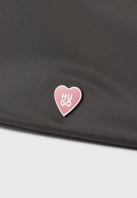 Ροζ καρφίτσα σε σχήμα καρδιάς με λευκό κείμενο "HUGO" σε σκούρο υφασμάτινο φόντο, πιθανόν μέρος τσάντας ή αξεσουάρ ρούχων.