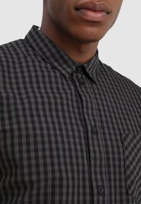 Czarna i szaro-kasztanowa koszula w kratę z krótkim rękawem, kołnierzykiem na guziki, zapinana z przodu na guziki i z kieszenią na piersi. Gładka tekstura tkaniny.