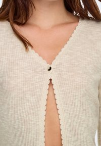 Gros plan sur un cardigan crème côtelé avec des bords festonnés, fermé par un petit bouton unique au niveau de la clavicule sur une personne aux cheveux bruns.