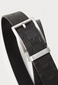 Cinturón de cuero negro con patrón geométrico embosado, hebilla de metal plateado y nombre de la marca grabado. Longitud ajustable con múltiples agujeros.